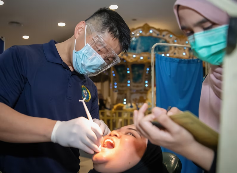 A male dentist is examining a patient's mouth while a female assistant taking notes