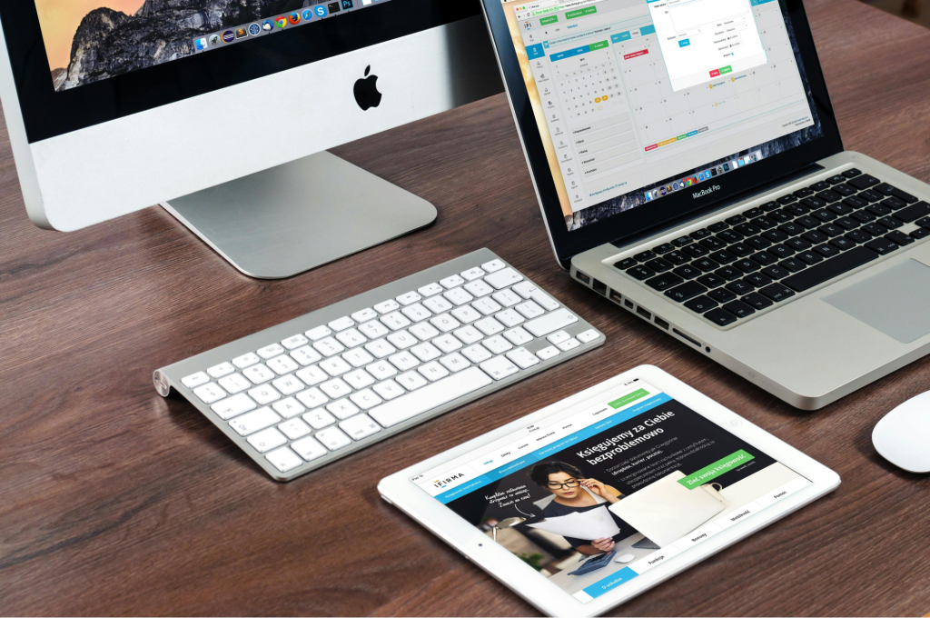 A desktop computer, a laptop, and a tablet are arranged on a wooden desk