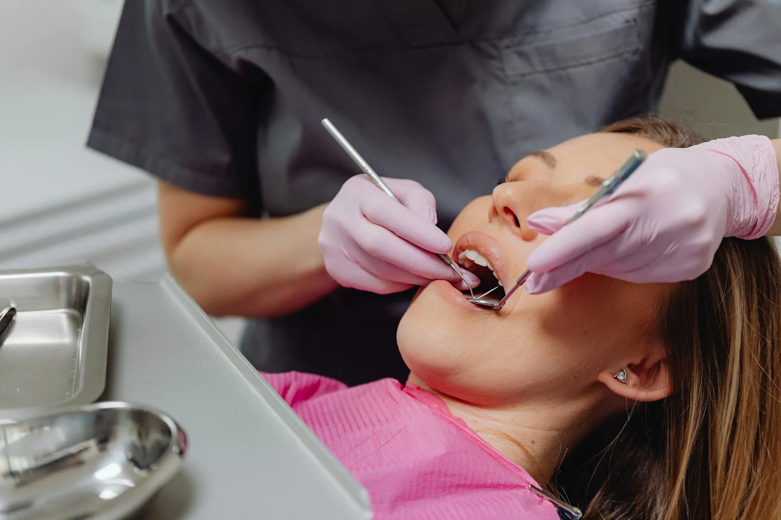 A dentist using dental tools to examine a female patient's open mouth.