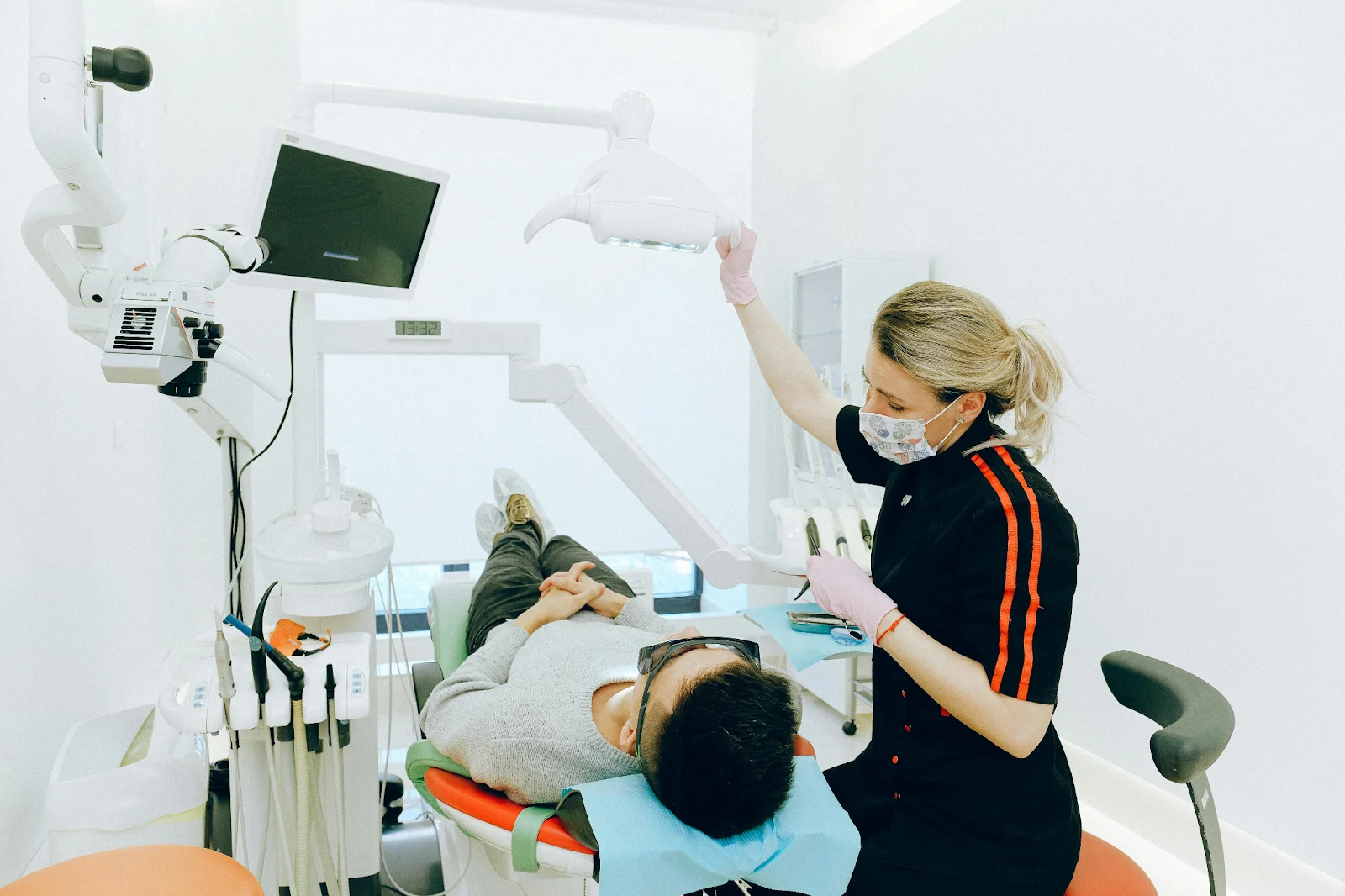 A dental professional wearing a mask and pink gloves is adjusting a dental lamp above a patient lying in a chair
