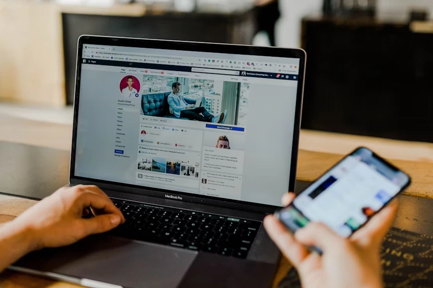 Hands operate a laptop showing a facebook profile while one hand holds a smartphone